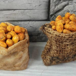 Op de afbeelding: Twee jute zakken gevuld met kleine, langwerpige, geel-oranje aardappelen. De zakken zijn gemaakt van een ruw, geweven materiaal. De achtergrond is een grijze stenen muur. De nummers 1 en 2 staan in blauwe vakken.