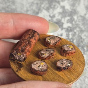Puede incluir: Miniatura de tabla de madera para servir con una salchicha a la parrilla y varias rodajas de salchicha. La salchicha es marrón con acentos rojo-anaranjados, y las rodajas revelan un relleno de carne con detalles amarillos y blancos. La tabla es de color marrón claro.