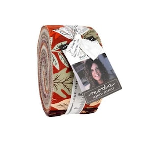 May include: A roll of fabric strips featuring a pattern of autumn leaves in shades of orange, red, and cream. A white ribbon secures the roll, attached to a rectangular tag with the text moda FABRICS + SUPPLIES and a photograph of a person.