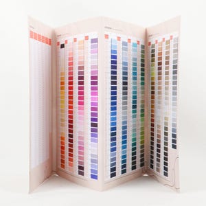 May include: A multi-page color chart fan book with a light pink cover. The pages display numerous vertical columns of thread color swatches, ranging from reds and yellows to blues and greens. The chart is open, revealing a wide array of hues.
