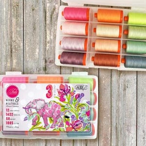 May include: A clear plastic storage box with 12 spools of Aurifil thread. The spools are arranged in two rows of six. The spools are in various shades of pink, orange, yellow, green, and blue. The box has a label that says "Neons & Neutrals" and "12 Small Spools 50 wt 100% Cotton".