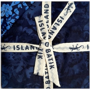 May include: Close-up of a gift wrapped in blue batik fabric, tied with a cream-colored ribbon. The ribbon is printed with the words "ISLAND BATIK" and features a lizard design. The fabric has a floral pattern.