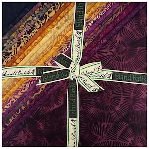 May include: A stack of batik fabric squares in shades of purple, gold, and brown, tied with a ribbon that reads "Island Batik". The fabric has various patterns, including swirls and abstract designs, and is neatly arranged.