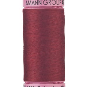 May include: A spool of dark burgundy sewing thread with light pink plastic ends. The top end features the text AMANN GROUP, and the bottom end displays the text 100% Polyester No. The thread is wound tightly around the central cylinder.