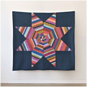 May include: A square quilt featuring a large, central eight-pointed star design. The star is composed of numerous thin, multicolored fabric strips in shades of pink, orange, yellow, blue, and purple, set against a solid dark navy blue background.