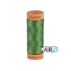 May include: A spool of green Aurifil thread. The thread is wound around a wooden spool with the brand name "AURIFIL" printed on the top and "MADE IN ITALY" on the bottom. The thread is a solid, medium green color.