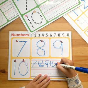 May include: A white dry-erase board with yellow borders and the word "Numbers" printed in red. The numbers 1 through 10 are printed in black above the board. The numbers 7, 8, 9, and 10 are written in blue on the board. A hand is writing the numbers 7, 8, 9, and 10 on the board with a blue marker.
