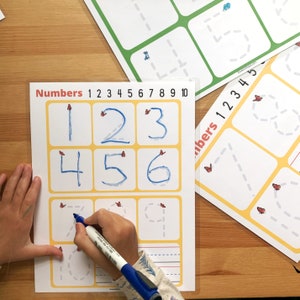 May include: A yellow and white worksheet with the word "Numbers" and the numbers 1 through 10 printed at the top. The numbers 1 through 6 are written in blue ink in the boxes below. A child's hand is tracing the dotted lines of the number 0 with a blue marker.