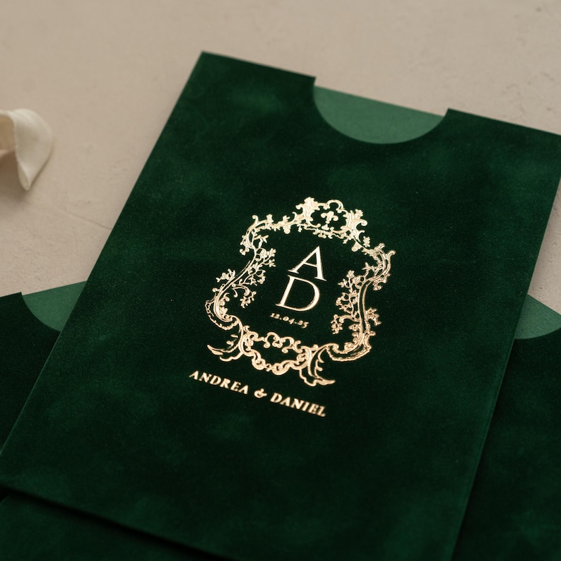 A close-up of a green velvet envelope with a gold embossed monogram and text on it.