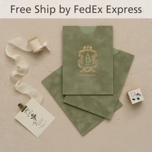 Może przedstawiać: Oliwkowe koperty zaproszeń ze złotymi literami i ozdobnym herbem. Mała karta ze słowem "merci" i kwiatową ilustracją jest przypięta do małego schowka. Obecna jest również szpula kremowej wstążki i stempel.