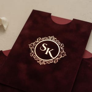 A close-up image of a burgundy-colored envelope or card with a gold-colored circular logo featuring the letters 'SK' in the center.