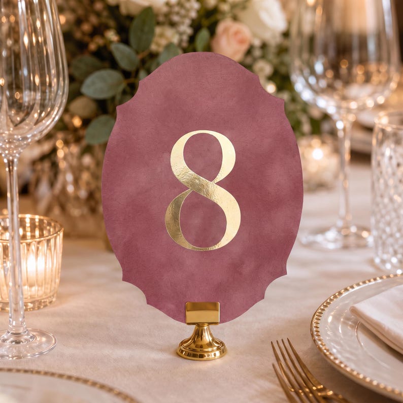 a table setting with a pink oval-shaped table number holder displaying the number 8, placed on a gold stand. The table is set with wine glasses, plates, and other tableware, creating an elegant and formal dining atmosphere.