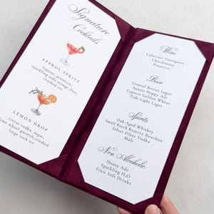 May include: A deep burgundy menu holder with two white inserts featuring printed text and watercolor illustrations of cocktails. The left side lists Signature Cocktails, Aperol Spritz, and Lemon Drop. The right side lists Wine, Beer, Spirits, and Non Alcoholic.
