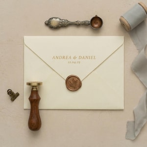 Op de afbeelding: Ivoorkleurige envelop verzegeld met een bruine wasstempel met een bladmotief. De envelop is bedrukt met de namen "Andrea & Daniel" en de datum "12.04.25" in goud. Een wasstempel met houten handvat, een metalen lepel en een lintspoel zijn ook aanwezig.