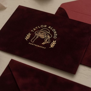 Può includere: Buste in velluto bordeaux con un logo dorato che recita "Taylor Alonso Photography". Il logo presenta un disegno a tratto di una persona che tiene una macchina fotografica, circondata da foglie. Le buste rettangolari hanno un interno bordeaux abbinato.