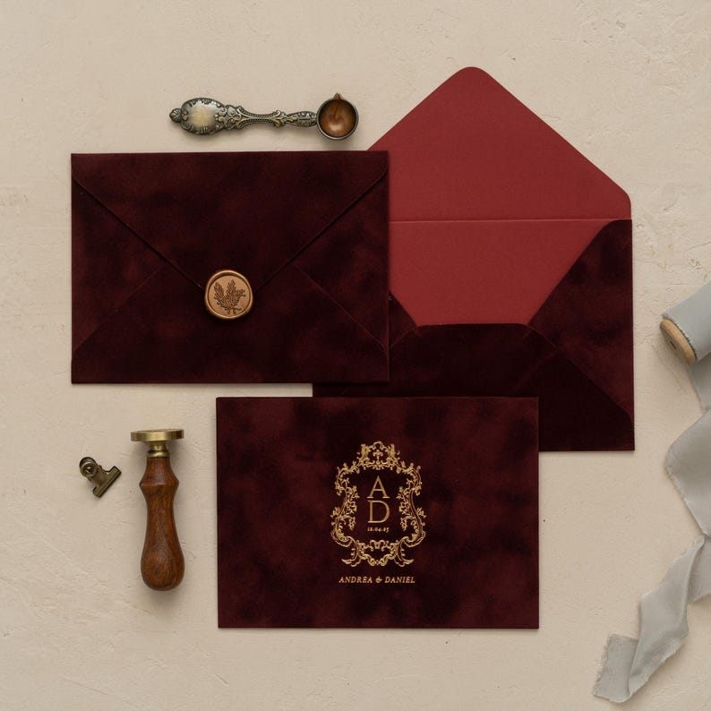 a burgundy envelope with a gold emblem, a red envelope, a wooden seal, and a silver spoon, all arranged on a white surface.