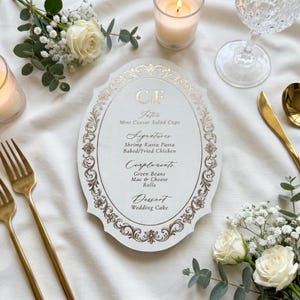 Könnte beinhalten: Eine ovale weiße Menükarte mit goldfarbenen Filigranrändern und goldener Schrift. Das Menü listet auf: CF, Intra Mini Ceasar Salad Cups, Signatures Shrimp Rasta Pasta Baked/Fried Chicken, Complements Green Beans Mac & Cheese Rolls, Dessert Wedding Cake.