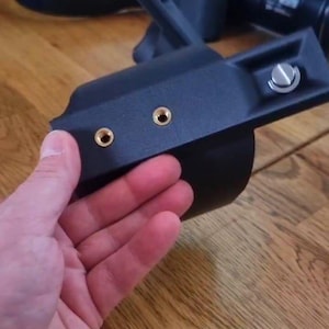 May include: A black camera accessory mount featuring a curved frame, two gold-coloured threaded mounting points, and a silver-toned screw mechanism. The device is constructed from a matte black material and includes an angled support arm.