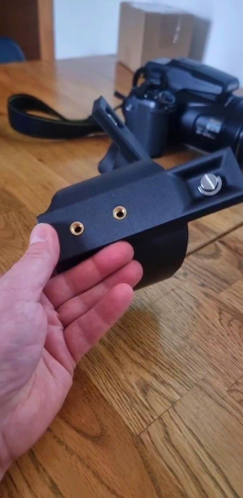 May include: A black camera accessory mount featuring a curved frame, two gold-coloured threaded mounting points, and a silver-toned screw mechanism. The device is constructed from a matte black material and includes an angled support arm.