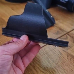May include: A black camera accessory, possibly a lens hood or a bracket, is held in a person's hand. The accessory has a rectangular shape with a curved top and a flat base. The background includes a camera and a wooden surface.