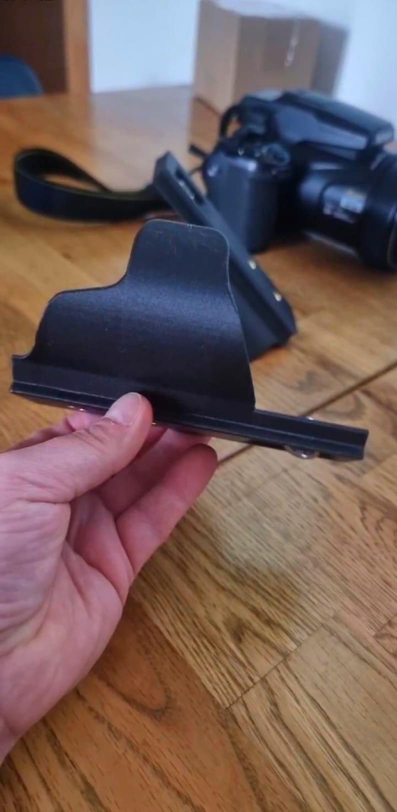 May include: A black camera accessory, possibly a lens hood or a bracket, is held in a person's hand. The accessory has a rectangular shape with a curved top and a flat base. The background includes a camera and a wooden surface.