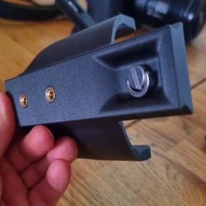 May include: A black camera accessory with a mounting plate, featuring two gold-coloured screw holes and a silver screw. The accessory is held in a hand, with a camera and other items in the background.