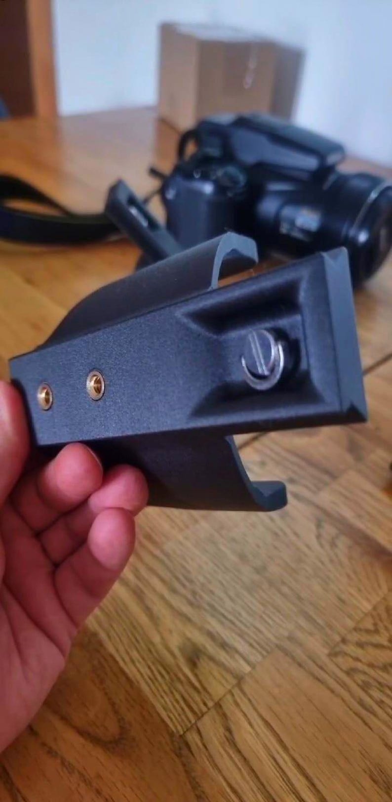 May include: A black camera accessory with a mounting plate, featuring two gold-coloured screw holes and a silver screw. The accessory is held in a hand, with a camera and other items in the background.