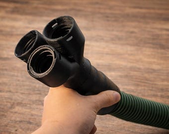 May include: A black plastic Y-shaped hose connector with three threaded openings. The connector is held in a hand and is attached to a green corrugated hose. The background is a wooden surface.