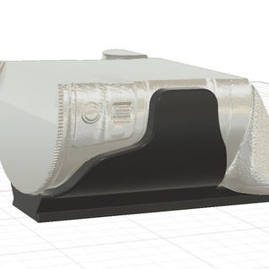 May include: A 3D-rendered, light grey object with a black interior and base. The object has a rounded, angular shape with textured details and a small, circular protrusion on top. The design includes a recessed area and a series of parallel lines.