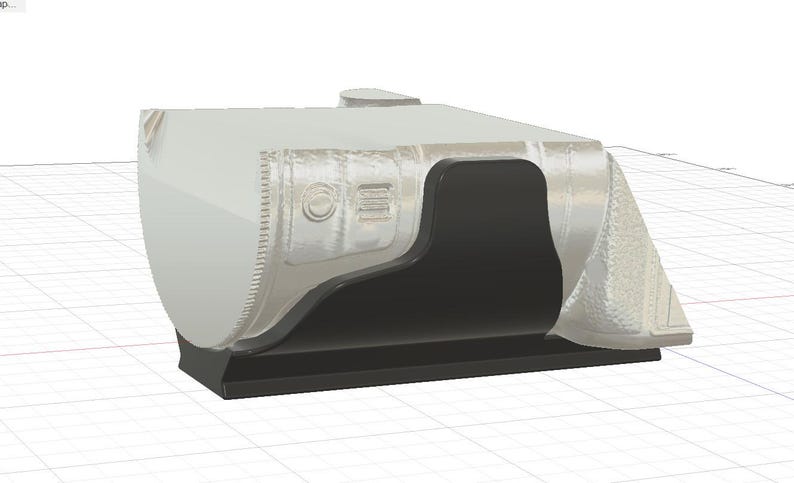May include: A 3D-rendered, light grey object with a black interior and base. The object has a rounded, angular shape with textured details and a small, circular protrusion on top. The design includes a recessed area and a series of parallel lines.