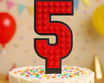 May include: A red number 5 cake topper with a black outline, designed to resemble a building block. The topper is placed on a white frosted cake decorated with colorful sprinkles. Balloons and party decorations are in the background.