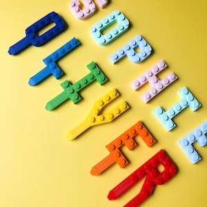 Könnte beinhalten: Bunte, 3D-gedruckte Buchstaben, die das Wort "SHOPPER" bilden, diagonal auf gelbem Hintergrund angeordnet. Die Buchstaben, in verschiedenen Farben wie Blau, Grün, Gelb, Orange und Rot, sind als Spielzeugbausteine gestaltet.