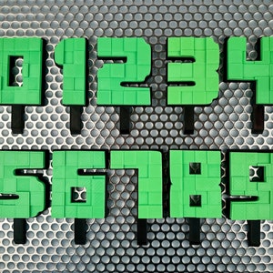 Può includere: Numeri di plastica verde da 0 a 9, ogni numero è su un bastoncino nero, i numeri sono disposti su due file, i numeri sono su uno sfondo argentato con un motivo circolare.