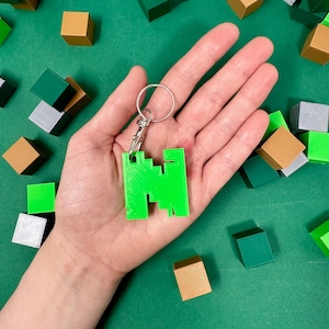 Könnte beinhalten: Ein grüner Plastik-Schlüsselanhänger in Form des Minecraft-Charakters Steve. Der Schlüsselanhänger ist an einem silbernen Metallring befestigt. Der Schlüsselanhänger ist von kleinen, bunten Blöcken umgeben.