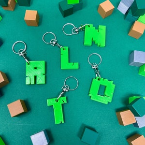 May include: A set of six green keychains shaped like letters, spelling out the word "ALOT". The keychains are on a green surface with brown and gray blocks.