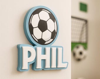 May include: A wall decoration with a soccer ball design, light blue border, and the name "PHIL" in white. The soccer ball is black and white. The background shows a soccer goal and additional soccer balls.