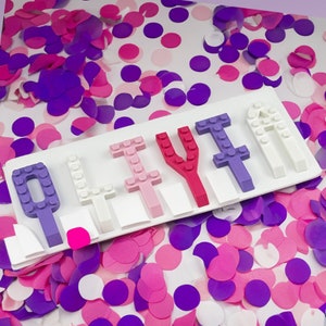 May include: A white tray with the word "LILY" spelled out in colorful plastic letters. The letters are arranged in a row on the tray, with the "L" on the left and the "Y" on the right. The tray is surrounded by pink and purple confetti.