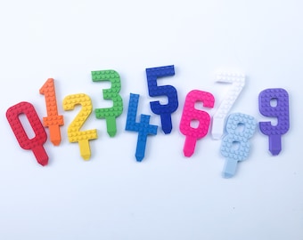 May include: A set of ten colorful plastic number candles, each with a different color and number from 0 to 9. The candles are arranged in a row from left to right, with the number 0 on the left and the number 9 on the right.