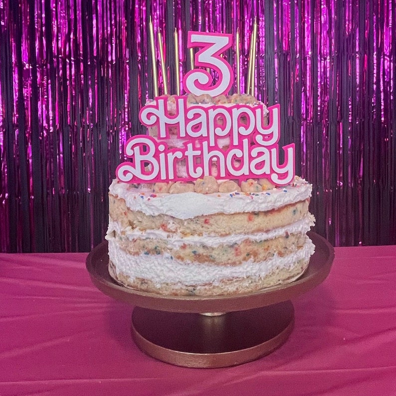 May include: A layered birthday cake with white frosting and colorful sprinkles, topped with a pink and white "Happy Birthday" sign and the number "3". The cake sits on a gold cake stand.