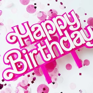 May include: A pink and white "Happy Birthday" cake topper with a decorative font. The topper is set against a white background with scattered pink confetti, creating a festive and celebratory scene.
