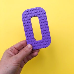 May include: A purple letter "O" with a studded surface, held against a bright yellow background. The letter is a solid color and has a rectangular shape with a hole in the middle. The studded texture gives it a playful, toy-like appearance.
