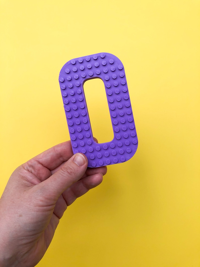 May include: A purple letter "O" with a studded surface, held against a bright yellow background. The letter is a solid color and has a rectangular shape with a hole in the middle. The studded texture gives it a playful, toy-like appearance.
