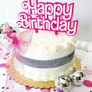 May include: A white frosted cake with a silver ribbon and a pink "Happy Birthday" cake topper. The cake sits on a gold scalloped cake board, with disco balls and pink confetti scattered around.