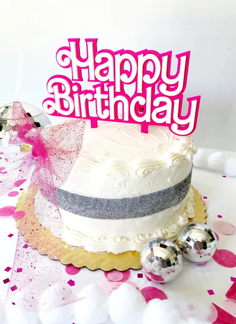 May include: A white frosted cake with a silver ribbon and a pink "Happy Birthday" cake topper. The cake sits on a gold scalloped cake board, with disco balls and pink confetti scattered around.