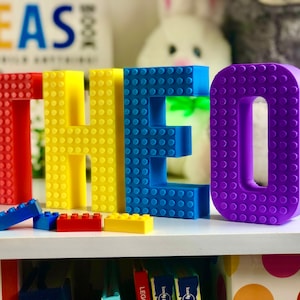 May include: A colorful display of the name "THEO" spelled out with large, 3D-printed building blocks in red, yellow, blue, and purple. Small building blocks in matching colors are scattered below. The background includes a shelf and a blurred book.