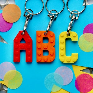 Może przedstawiać: Trzy srebrne breloki z kolorowymi plastikowymi zawieszkami w kształcie liter, które tworzą napis "ABC". Zawieszki są wykonane tak, aby przypominały klocki Lego. Breloki znajdują się na niebieskim tle z kolorowymi papierowymi kółkami.