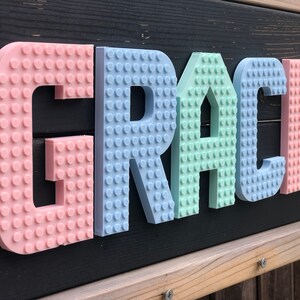 May include: A colorful sign spelling "GRACE" in pastel-colored, 3D-printed letters. The letters are designed to resemble interlocking toy bricks, with a pink "G", a light blue "R", a mint green "A", a light blue "C", and a pink "E".