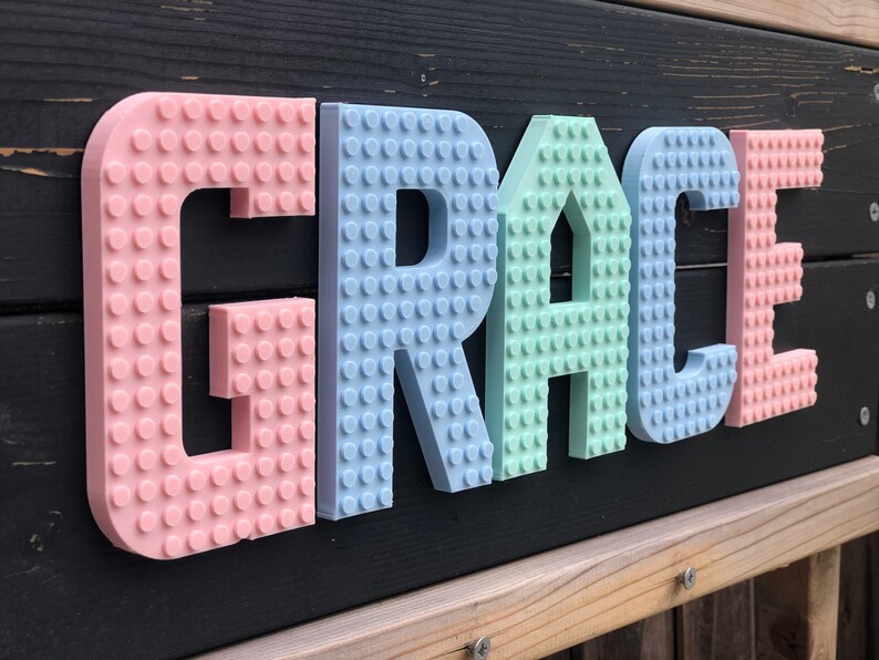 May include: A colorful sign spelling "GRACE" in pastel-colored, 3D-printed letters. The letters are designed to resemble interlocking toy bricks, with a pink "G", a light blue "R", a mint green "A", a light blue "C", and a pink "E".