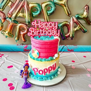 May include: A two-tiered birthday cake with pink frosting and blue and white accents. The cake is decorated with a beach ball and the name "Poppy". A "Happy Birthday" cake topper and a mermaid doll are also present.