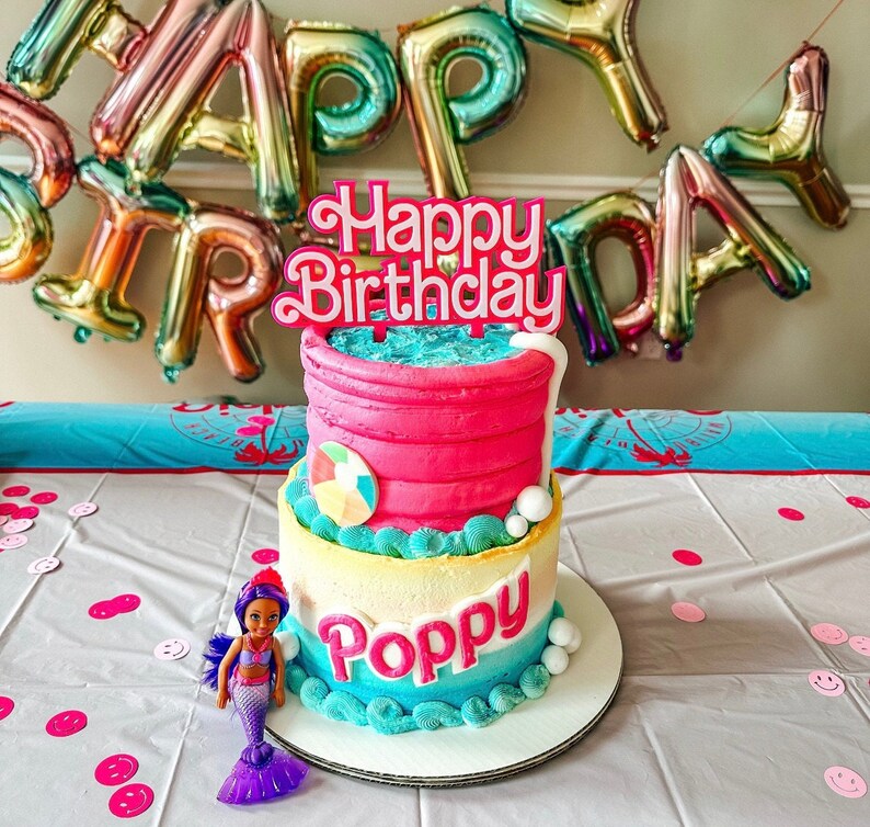 May include: A two-tiered birthday cake with pink frosting and blue and white accents. The cake is decorated with a beach ball and the name "Poppy". A "Happy Birthday" cake topper and a mermaid doll are also present.